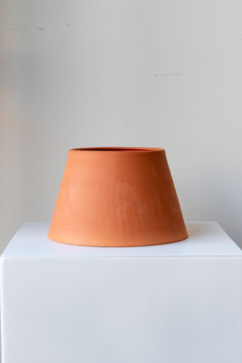 One terra cotta planter sits on a white surface in a white room. It is round and angled. It is wider at the base and more narrow at the top. The planter is empty and photographed straight on.