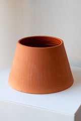 One terra cotta planter sits on a white surface in a white room. It is round and angled. It is wider at the base and more narrow at the top. The planter is empty and photographed at a slight angle to show the clay detail.