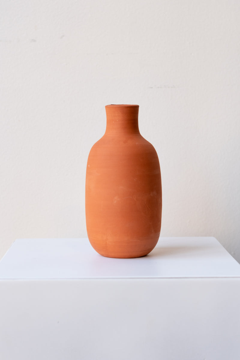 One terra cotta bud vase sits on a white surface in a white room. The vase is round and shaped like a bottle. The vase is empty. It is photographed straight on.