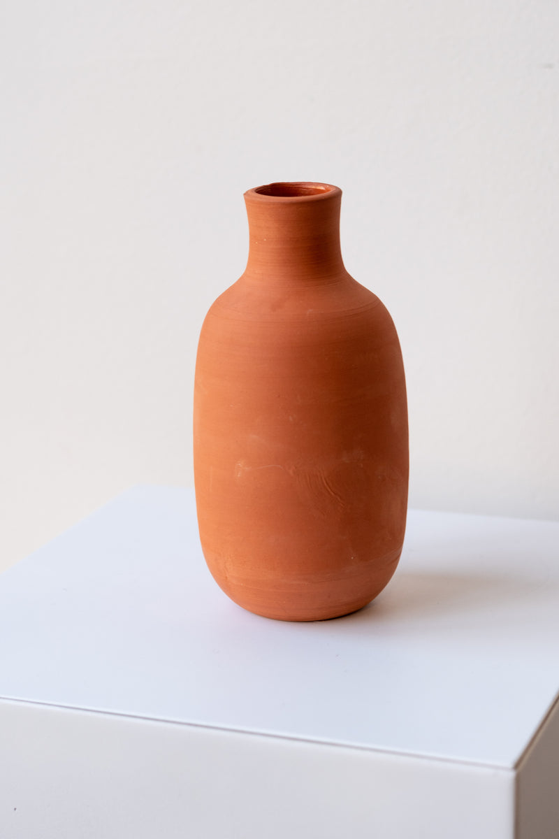 One terra cotta bud vase sits on a white surface in a white room. The vase is round and shaped like a bottle. The vase is empty. It is photographed closer and at a slight angle to show the clay details.