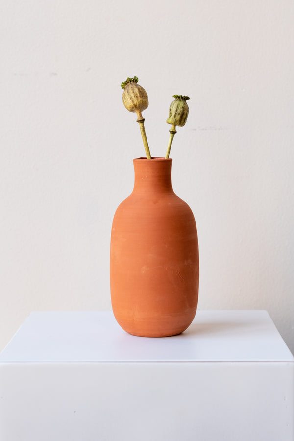 One terra cotta bud vase sits on a white surface in a white room. The vase is round and shaped like a bottle. There are two dried green poppy stems in the vase. It is photographed straight on.