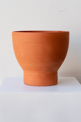One terra cotta vase sits on a white surface in a white room. The vase is cylindrical at the bottom with a wider bowl shaped top. The vase is empty. It is photographed straight on.