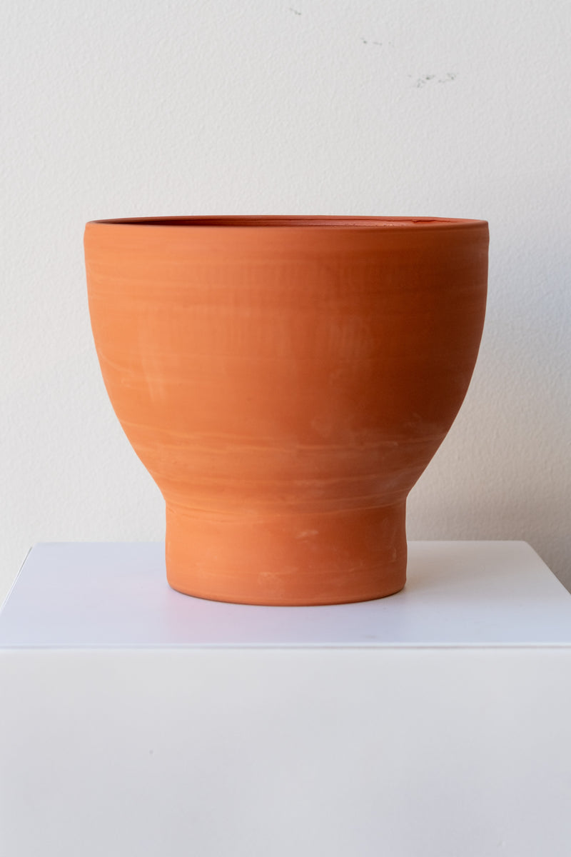 One terra cotta vase sits on a white surface in a white room. The vase is cylindrical at the bottom with a wider bowl shaped top. The vase is empty. It is photographed straight on.