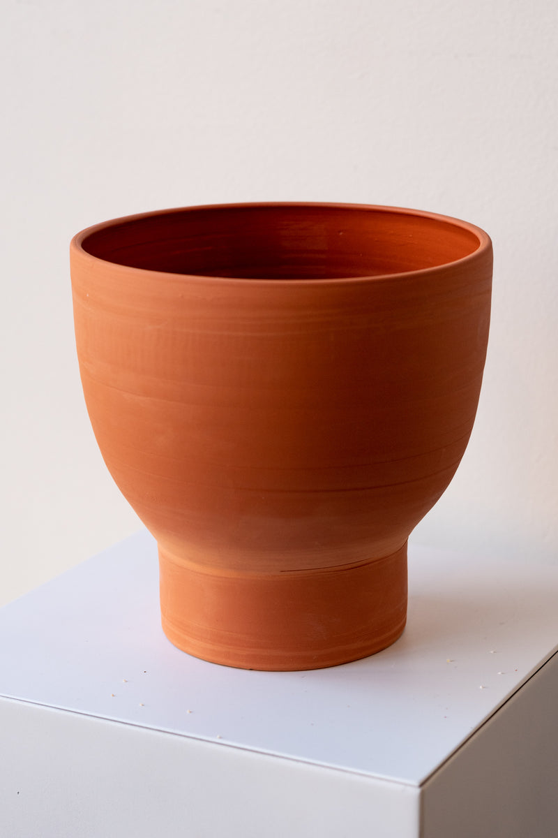 One terra cotta vase sits on a white surface in a white room. The vase is cylindrical at the bottom with a wider bowl shaped top. The vase is empty. It is photographed closer and at an angle.