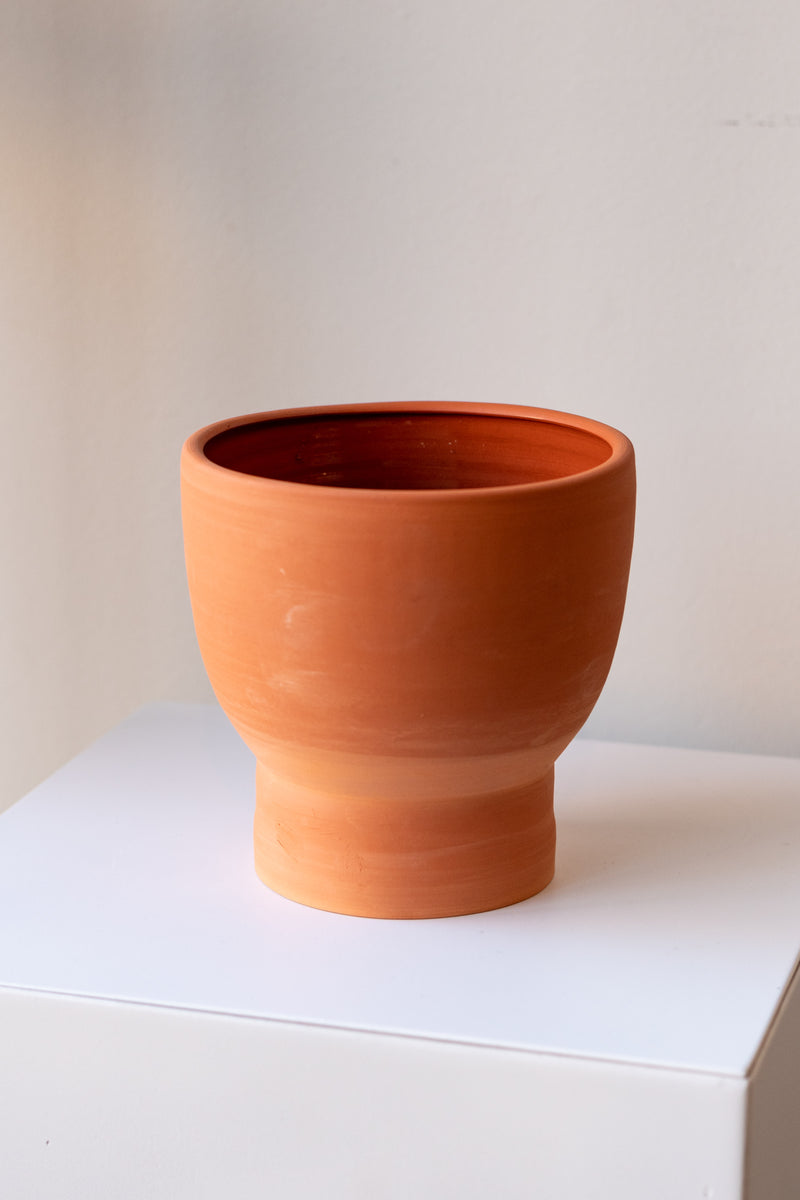 One terra cotta vase sits on a white surface in a white room. The vase is cylindrical at the bottom with a wider bowl shaped top. The vase is empty. It is photographed closer and at an angle.