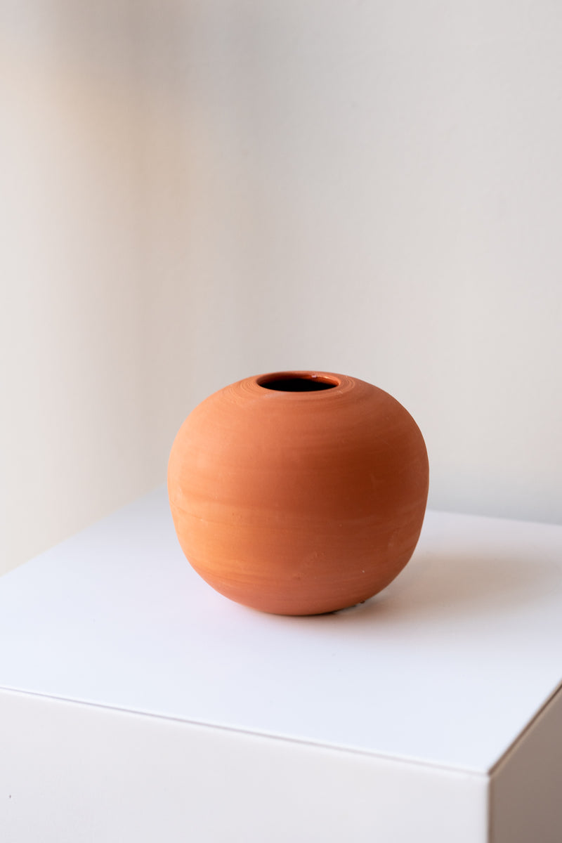 One small round terra cotta vase sits on a white surface in a white room. There are stems of small orange flowers in the vase. It is photographed closer and at an angle.