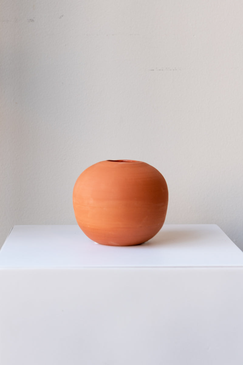 One small round terra cotta vase sits on a white surface in a white room. The vase is empty. It is photographed straight on.