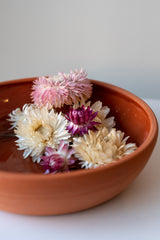 One medium terra cotta bowl sits on a white surface in a white room. The outside of the bowl is matte and unglazed, and the inside is glazed and shiny. The bowl has dried pink and white flowers in it.. It is photographed close up, and part of the bowl is out of the frame.
