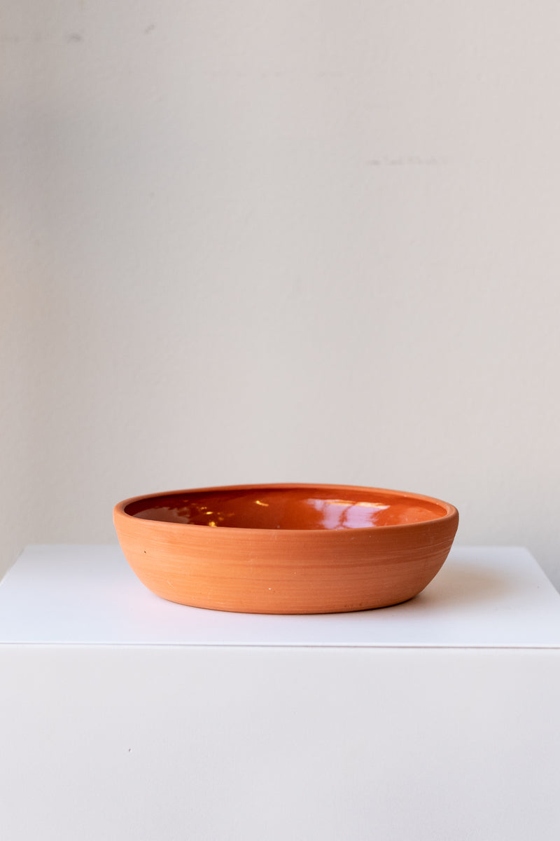 One medium terra cotta bowl sits on a white surface in a white room. The outside of the bowl is matte and unglazed, and the inside is glazed and shiny. The bowl is empty. It is photographed straight on.
