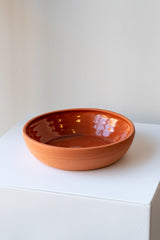 One medium terra cotta bowl sits on a white surface in a white room. The outside of the bowl is matte and unglazed, and the inside is glazed and shiny. The bowl is empty. It is photographed closer and at an angle.