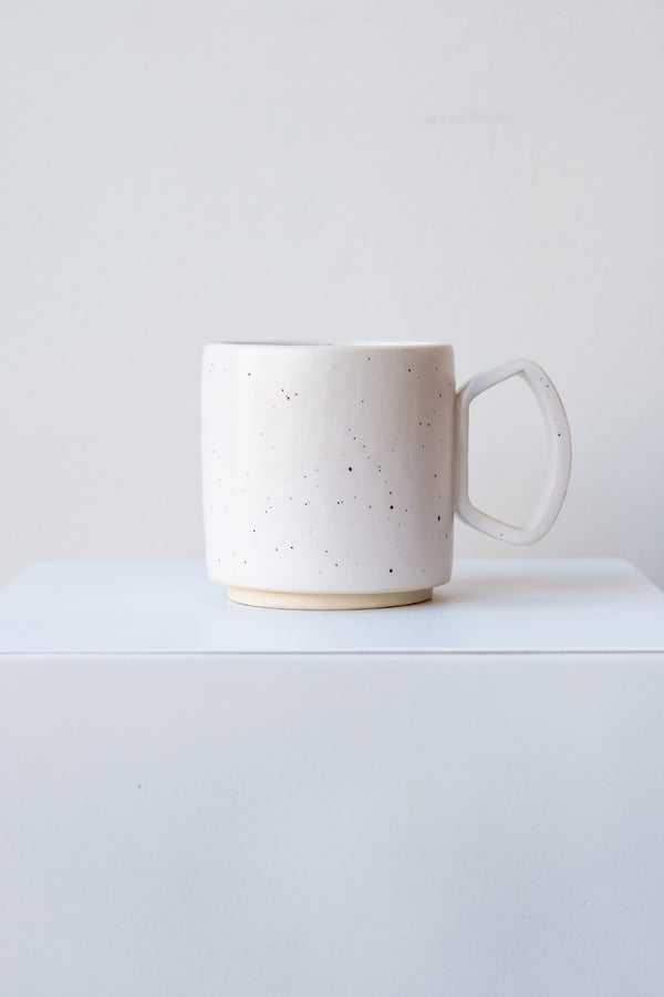 One ceramic mug sits on a white surface in a white room. The mug is white with black speckles. There is a narrow ring of unglazed clay at the bottom of the mug. It is photographed straight on.