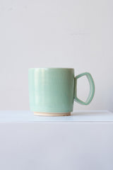 One ceramic mug sits on a white surface in a white room. The mug is a light blue-green. There is a narrow ring of unglazed clay at the bottom of the mug. It is photographed straight on.
