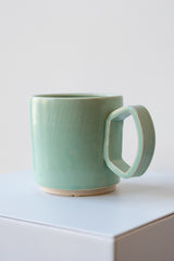One ceramic mug sits on a white surface in a white room. The mug is a light blue-green. There is a narrow ring of unglazed clay at the bottom of the mug. It is photographed at an angle to highlight the shape of the handle, which comes out from the cup at two angles joined by a curve.