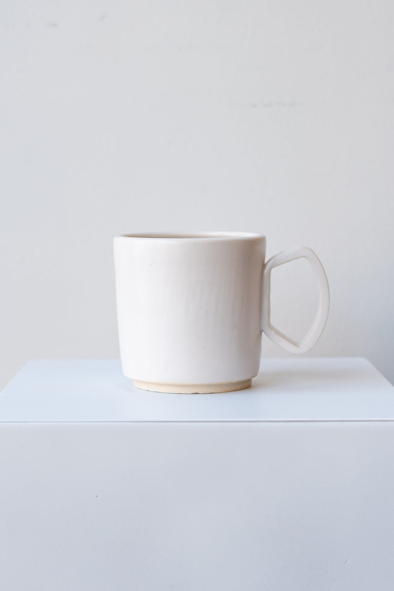 One ceramic mug sits on a white surface in a white room. The mug is white. There is a narrow ring of unglazed clay at the bottom of the mug. It is photographed straight on.