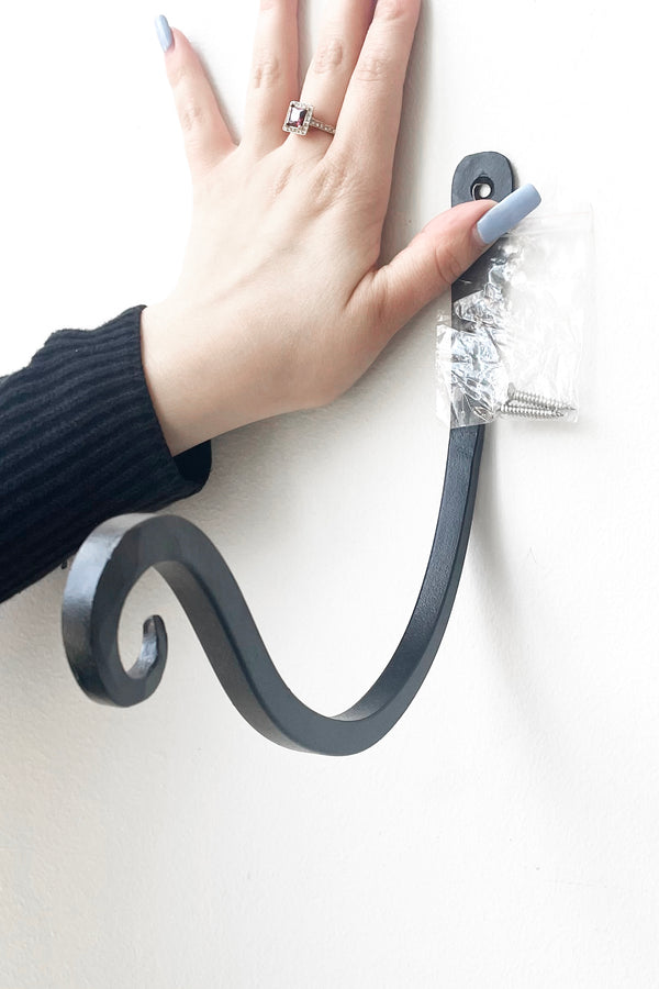 A hand holds Upcurled Metal Wallhook 8" against white wall