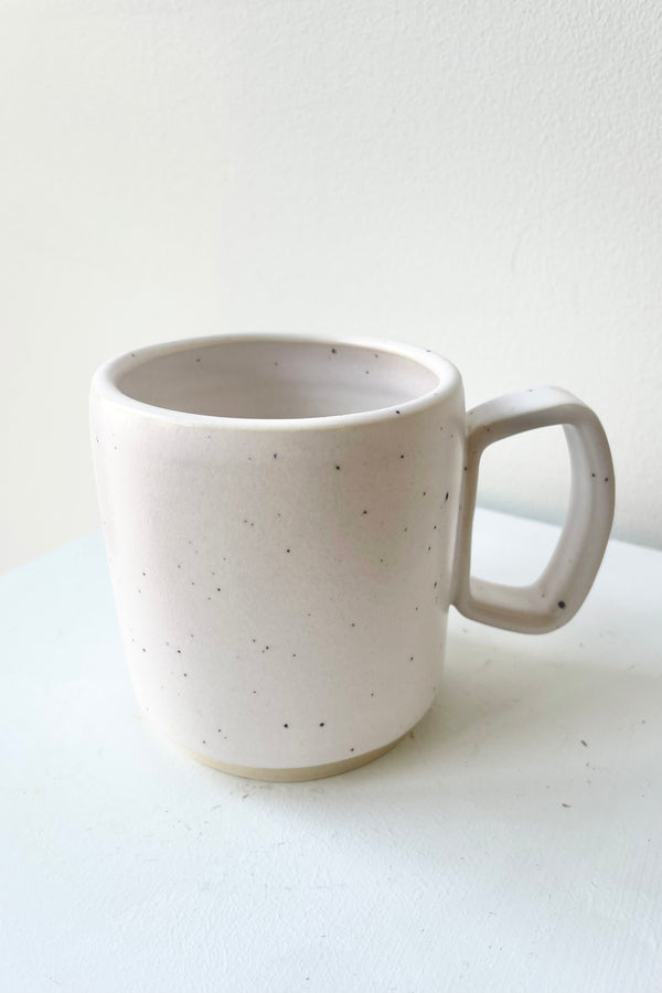 Detail of One ceramic mug sits on a white surface in a white room. The mug is white with black speckles. There is a narrow ring of unglazed clay at the bottom of the mug. It is photographed straight on.