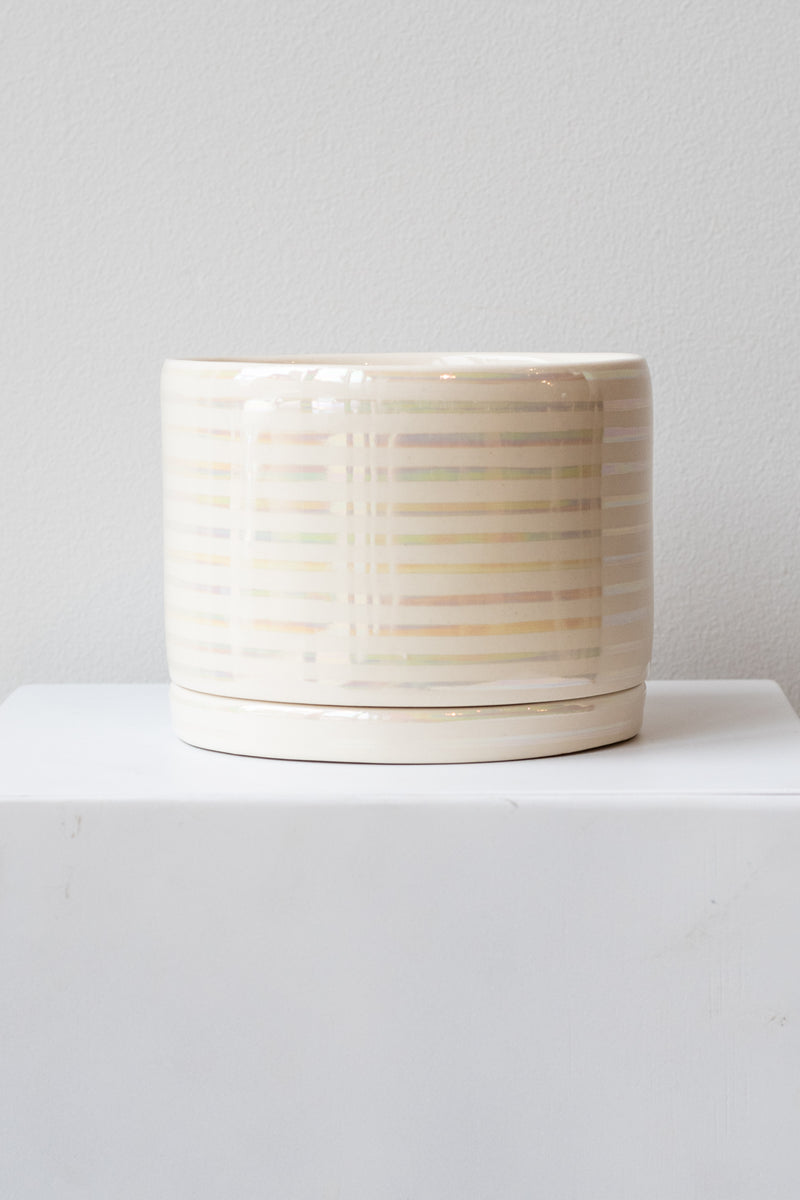 A large white ceramic planter sits on a white surface in a white room. The planter has thin stripes of iridescent glaze and a matching drainage tray. The planter is empty. It is photographed straight on.