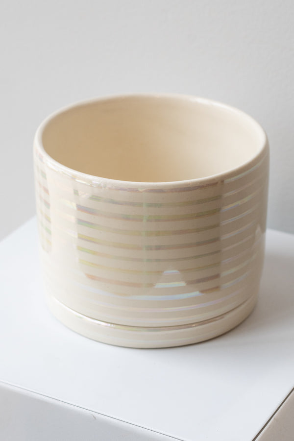A large white ceramic planter sits on a white surface in a white room. The planter has thin stripes of iridescent glaze and a matching drainage tray. The planter is empty. It is photographed closer and at an angle.