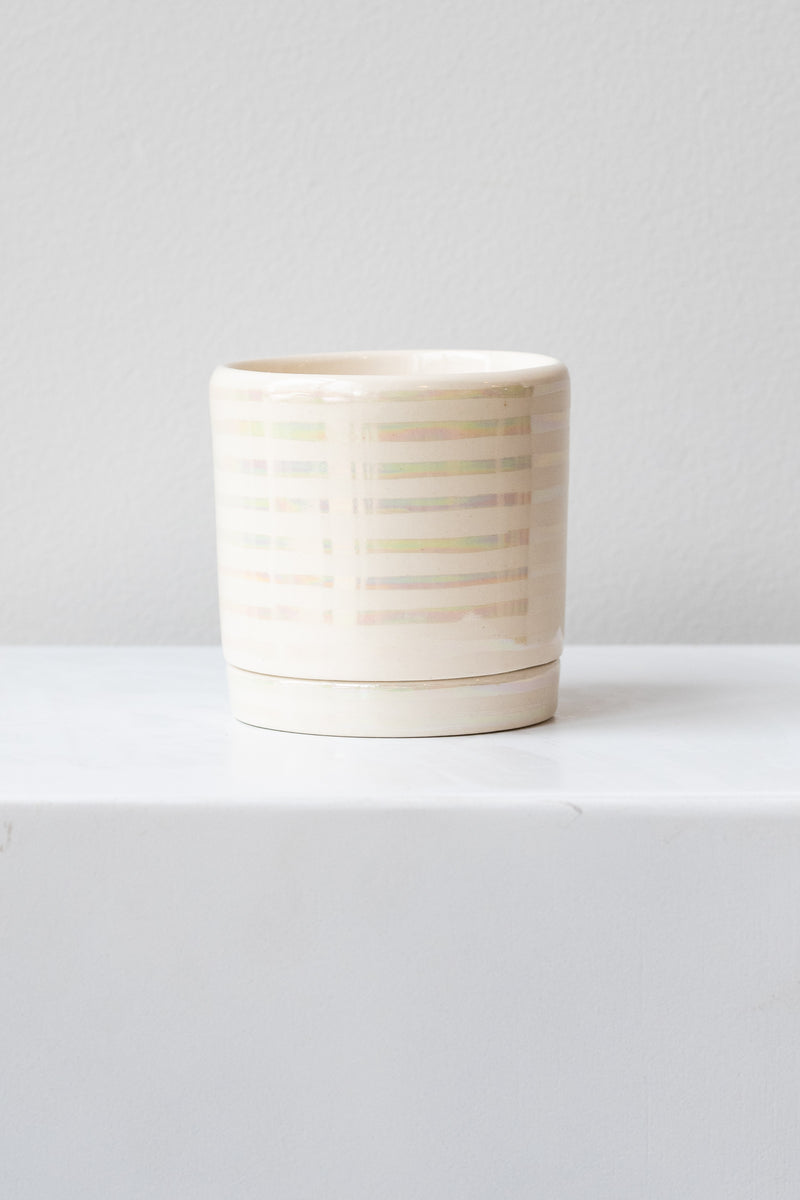 A small white ceramic planter sits on a white surface in a white room. The planter has thin stripes of iridescent glaze and a matching drainage tray. The planter is empty. It is photographed straight on.