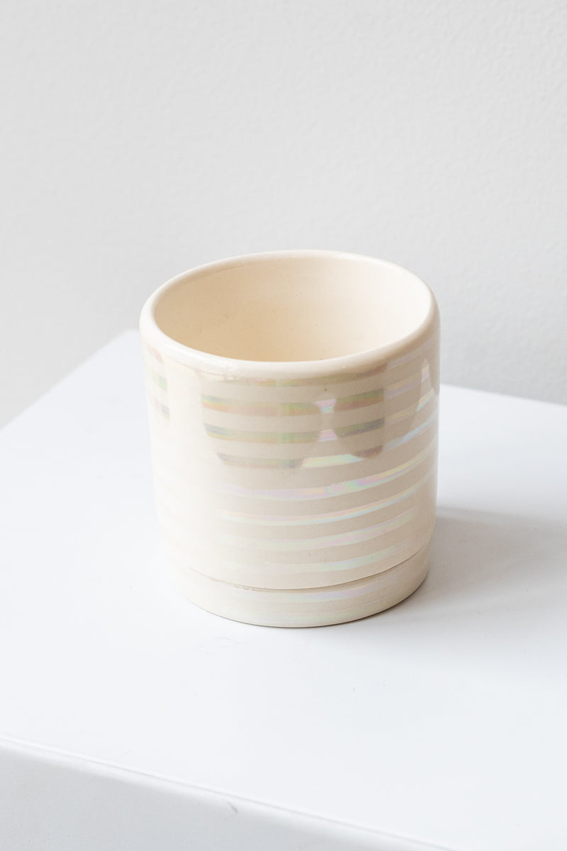 A small white ceramic planter sits on a white surface in a white room. The planter has thin stripes of iridescent glaze and a matching drainage tray. The planter is empty. It is photographed closer and at an angle.