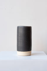 One small cylindrical clay vase sits on a white surface in a white room. The vase is glazed with a black glaze with white speckles. The bottom quarter of the vase is unglazed, showing cream-colored clay. The vase is empty. It is photographed straight on.
