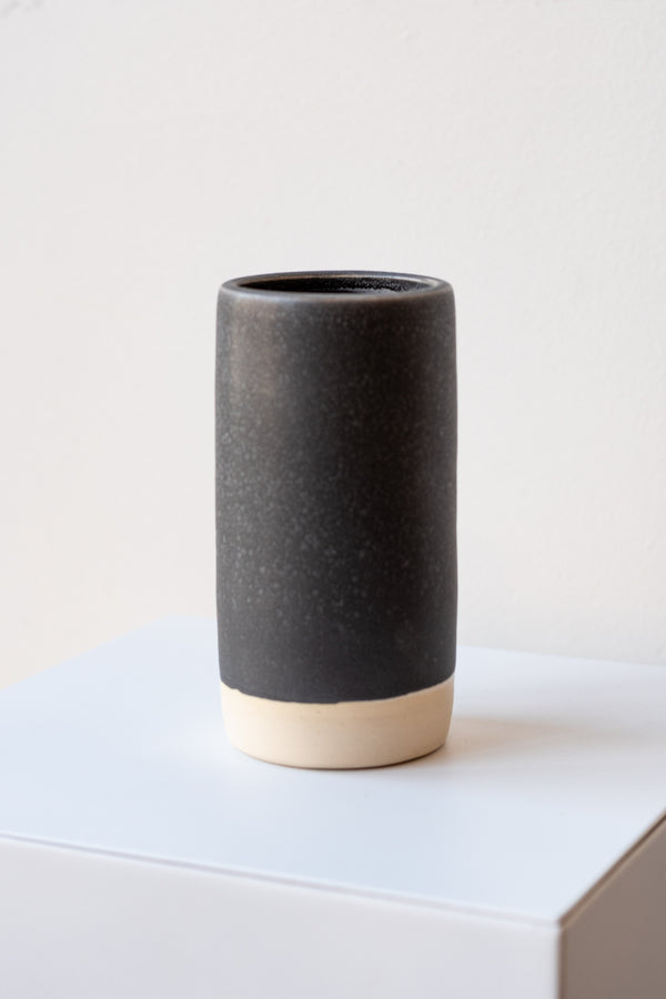 One small cylindrical clay vase sits on a white surface in a white room. The vase is glazed with a black glaze with white speckles. The bottom quarter of the vase is unglazed, showing cream-colored clay. The vase is empty. It is photographed closer and at an angle.