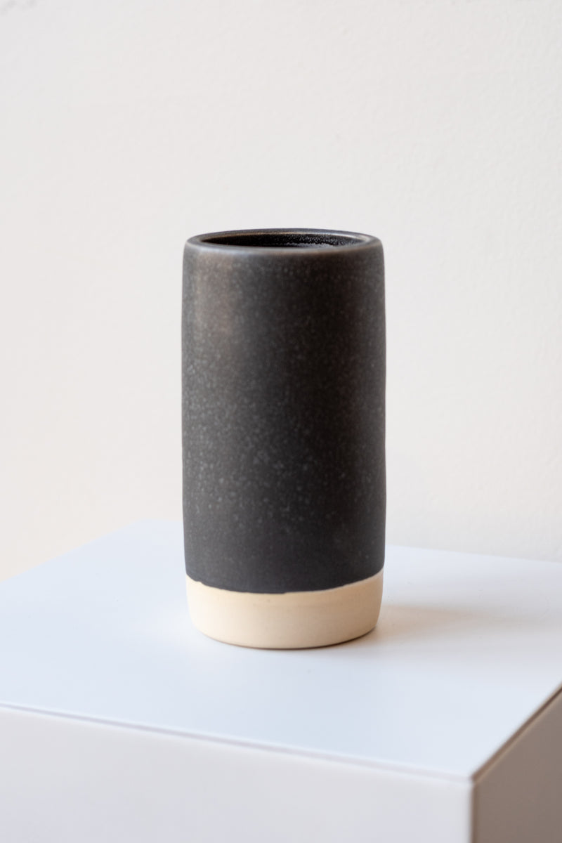 One small cylindrical clay vase sits on a white surface in a white room. The vase is glazed with a black glaze with white speckles. The bottom quarter of the vase is unglazed, showing cream-colored clay. The vase is empty. It is photographed closer and at an angle.