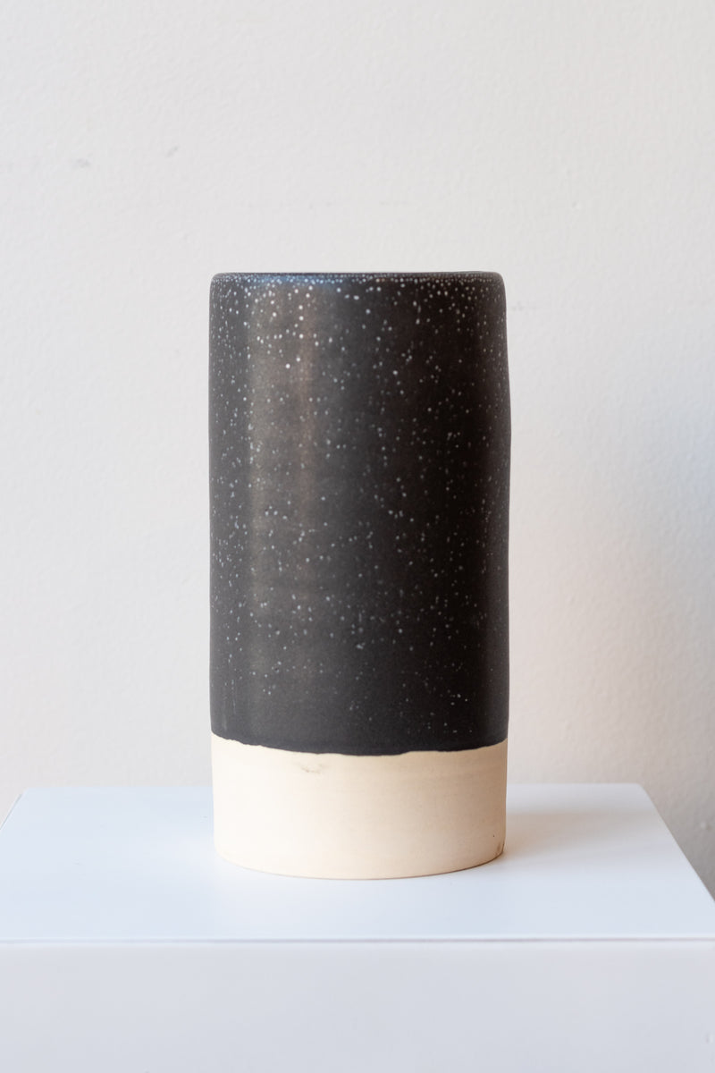 One cylindrical clay vase sits on a white surface in a white room. The vase is glazed with a black glaze with white speckles. The bottom quarter of the vase is unglazed, showing cream-colored clay. The vase is empty. It is photographed straight on.
