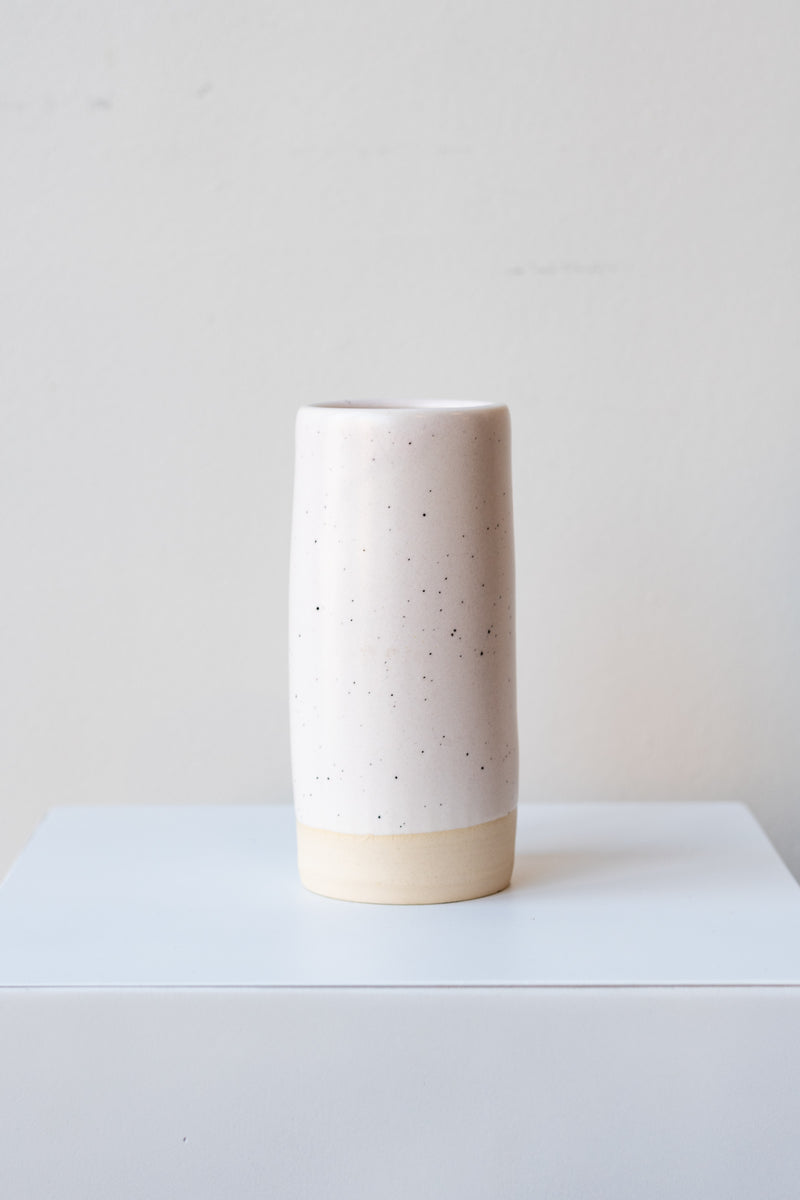 One small cylindrical clay vase sits on a white surface in a white room. The vase is glazed with a white glaze with black speckles. The bottom quarter of the vase is unglazed, showing cream-colored clay. The vase is empty. It is photographed straight on.