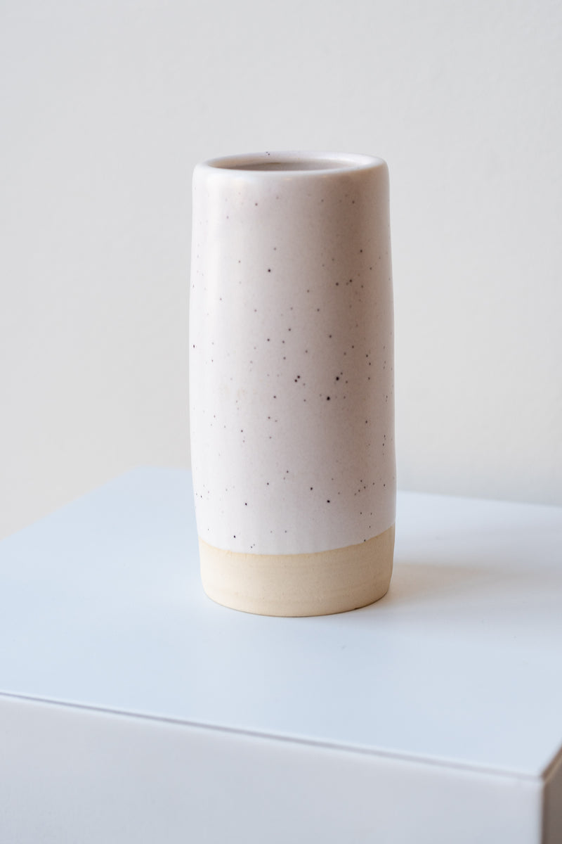 One small cylindrical clay vase sits on a white surface in a white room. The vase is glazed with a white glaze with black speckles. The bottom quarter of the vase is unglazed, showing cream-colored clay. The vase is empty. It is photographed closer and at an angle.