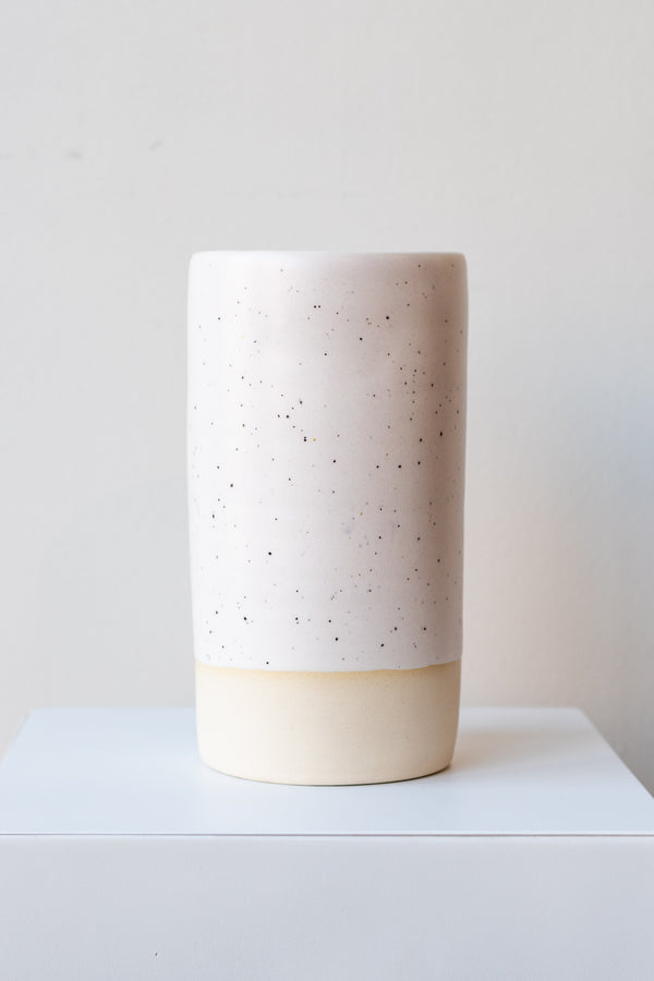 One cylindrical clay vase sits on a white surface in a white room. The vase is glazed with a white glaze with black speckles. The bottom quarter of the vase is unglazed, showing cream-colored clay. The vase is empty. It is photographed straight on.