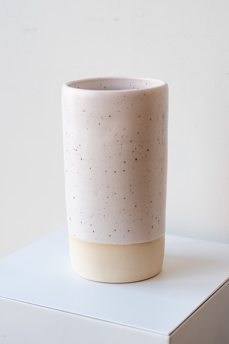 One cylindrical clay vase sits on a white surface in a white room. The vase is glazed with a white glaze with black speckles. The bottom quarter of the vase is unglazed, showing cream-colored clay. The vase is empty. It is photographed closer and at an angle.