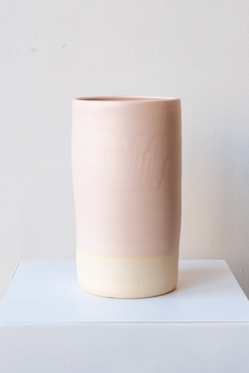 One cylindrical clay vase sits on a white surface in a white room. The vase is glazed with a light pink glaze. The bottom quarter of the vase is unglazed, showing cream-colored clay. The vase is empty. It is photographed straight on.