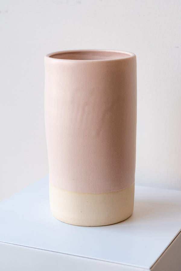 One cylindrical clay vase sits on a white surface in a white room. The vase is glazed with a light pink glaze. The bottom quarter of the vase is unglazed, showing cream-colored clay. The vase is empty. It is photographed closer and at an angle.