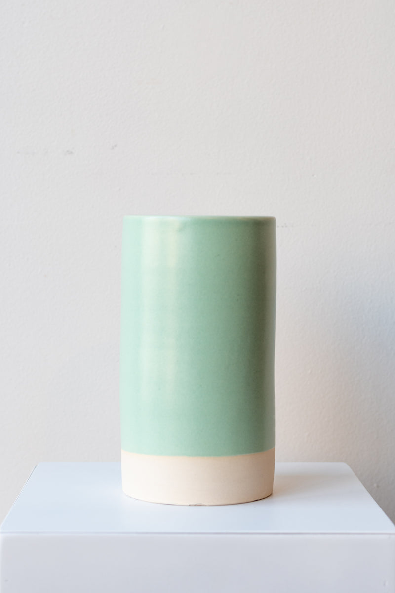 One cylindrical clay vase sits on a white surface in a white room. The vase is glazed with a light blue-green glaze. The bottom quarter of the vase is unglazed, showing cream-colored clay. The vase is empty. It is photographed straight on.