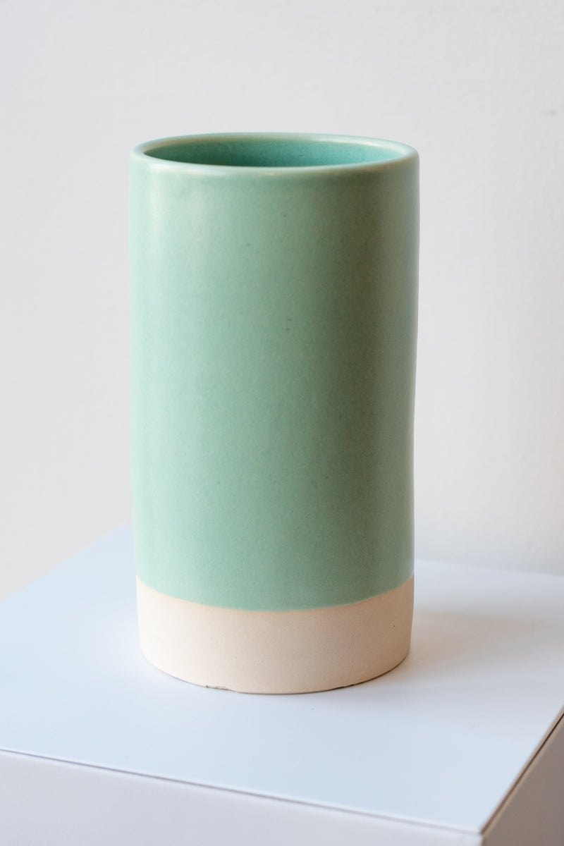 One cylindrical clay vase sits on a white surface in a white room. The vase is glazed with a light blue-green glaze. The bottom quarter of the vase is unglazed, showing cream-colored clay. The vase is empty. It is photographed closer and at an angle.