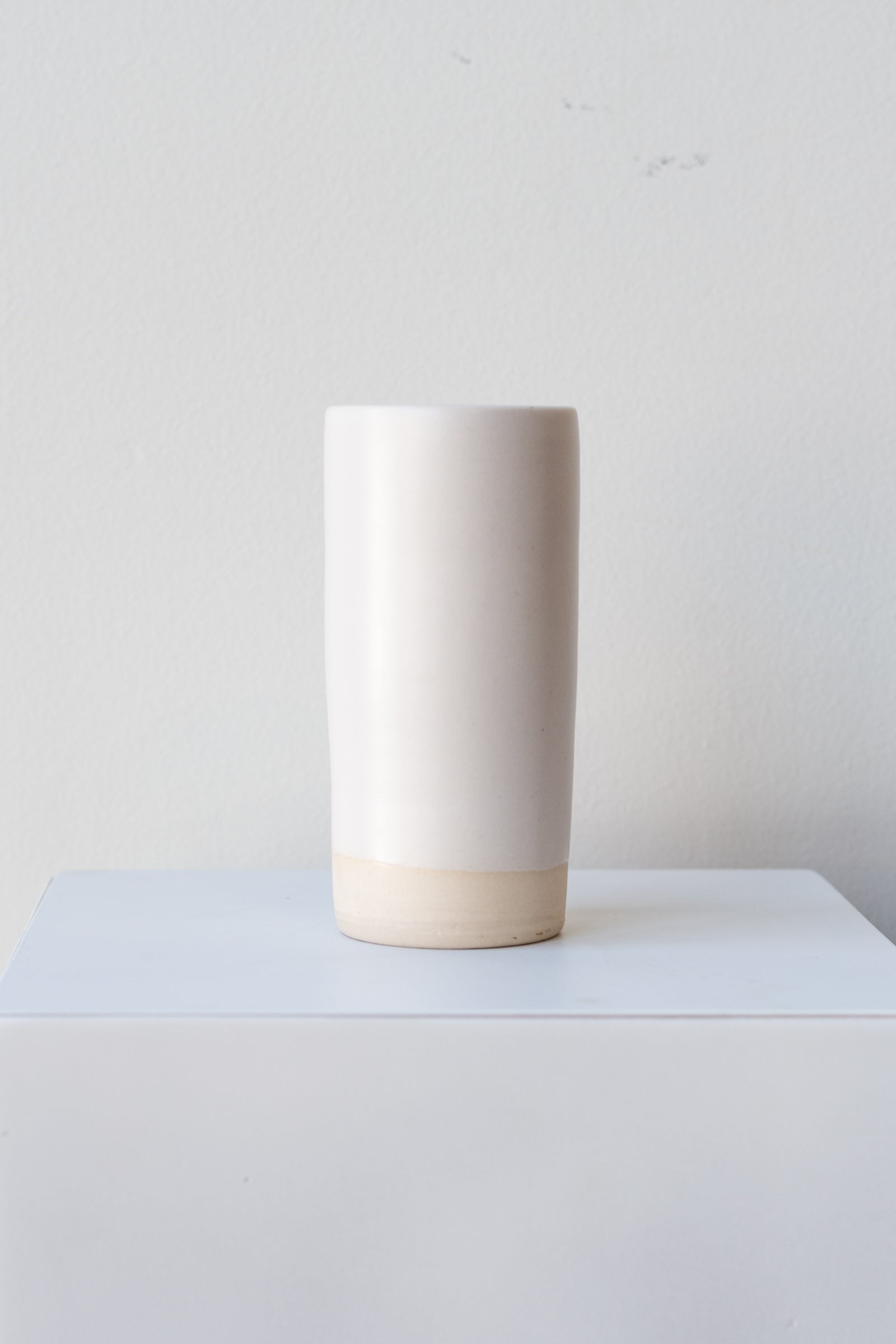 One small cylindrical clay vase sits on a white surface in a white room. The vase is glazed with a white glaze. The bottom quarter of the vase is unglazed, showing cream-colored clay. The vase is empty. It is photographed straight on.