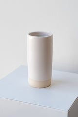 One small cylindrical clay vase sits on a white surface in a white room. The vase is glazed with a white glaze. The bottom quarter of the vase is unglazed, showing cream-colored clay. The vase is empty. It is photographed closer and at an angle.