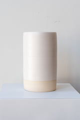 One cylindrical clay vase sits on a white surface in a white room. The vase is glazed with a white glaze. The bottom quarter of the vase is unglazed, showing cream-colored clay. The vase is empty. It is photographed straight on.