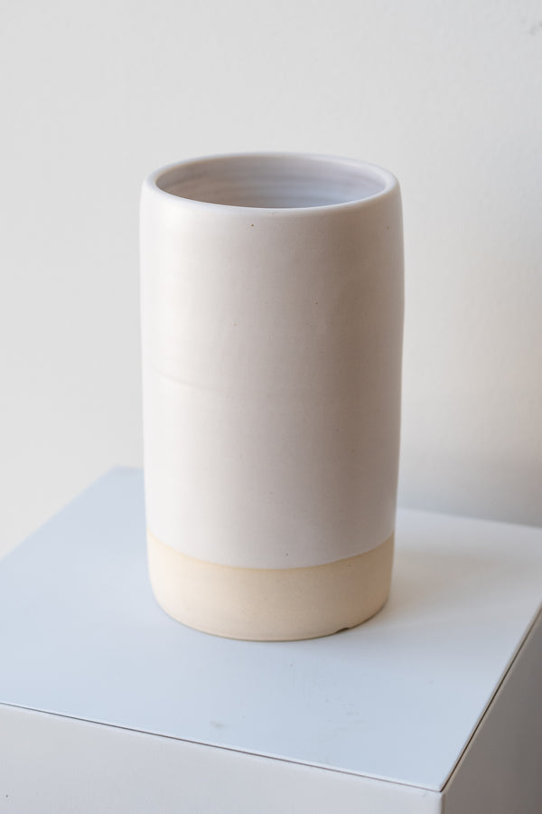 One cylindrical clay vase sits on a white surface in a white room. The vase is glazed with a white glaze. The bottom quarter of the vase is unglazed, showing cream-colored clay. The vase is empty. It is photographed closer and at an angle.
