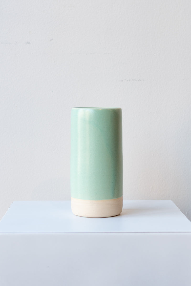 One small cylindrical clay vase sits on a white surface in a white room. The vase is glazed with a light blue-green glaze. The bottom quarter of the vase is unglazed, showing cream-colored clay. The vase is empty. It is photographed straight on.