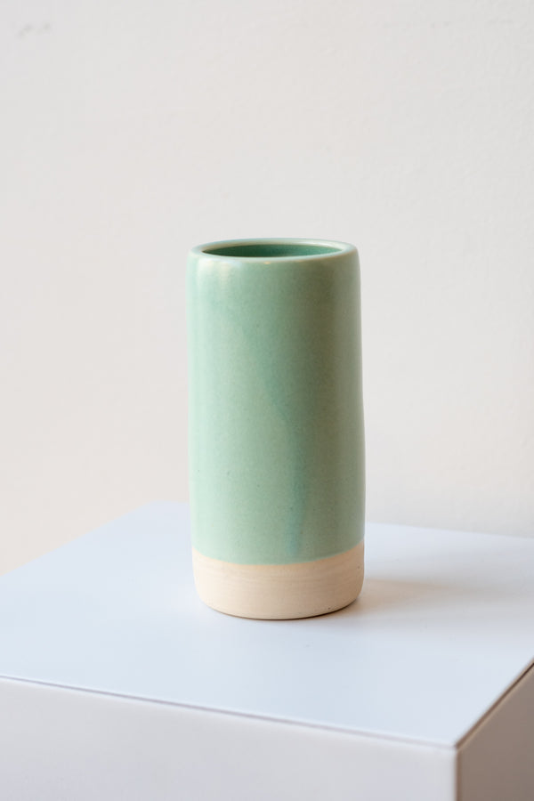 One small cylindrical clay vase sits on a white surface in a white room. The vase is glazed with a light blue-green glaze. The bottom quarter of the vase is unglazed, showing cream-colored clay. The vase is empty. It is photographed closer and at an angle.