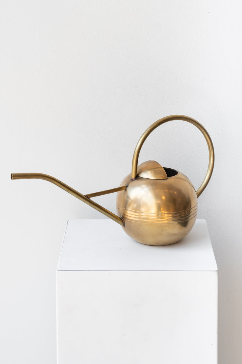 Round brass watering can with long thin spout and round handle sits on a white pedestal in a white room.