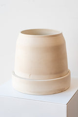 One cream colored stoneware planter sits on a white surface in a white room. The planter is round and squat, with a small logo imprinted in the clay on the bottom. The planter also sits on a round drainage tray. It is photographed at a slight angle to show the clay detail.