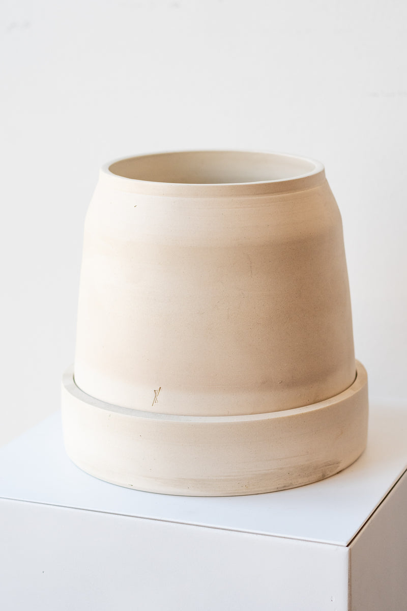 One cream colored stoneware planter sits on a white surface in a white room. The planter is round and squat, with a small logo imprinted in the clay on the bottom. The planter also sits on a round drainage tray. It is photographed at a slight angle to show the clay detail.