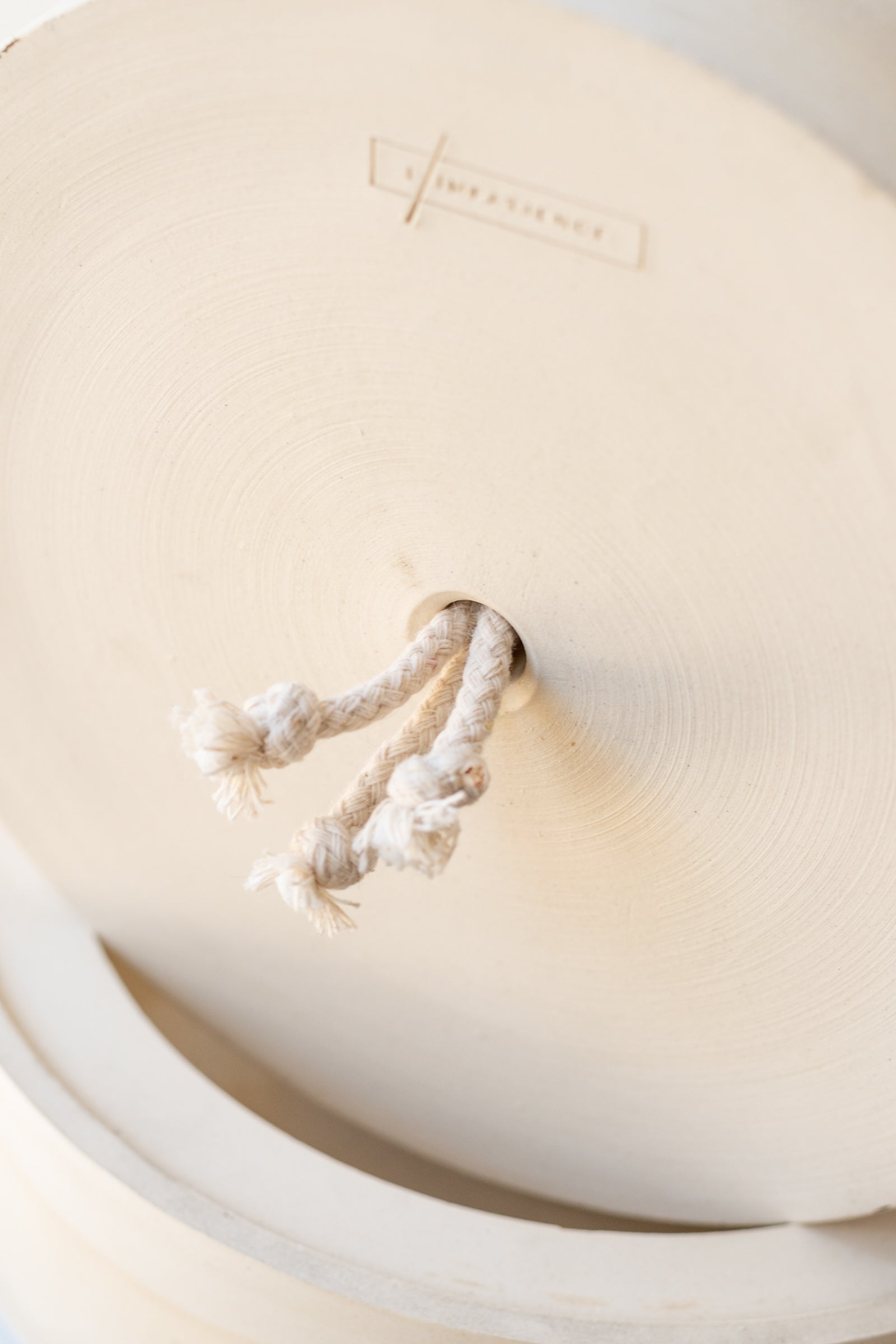 The bottom of a cream colored stoneware planter is tipped to show a hole on the bottom with three cotton wicks coming through it. It is resting on a round drainage tray of the same color. A logo for L'Impatience is etched into the clay.