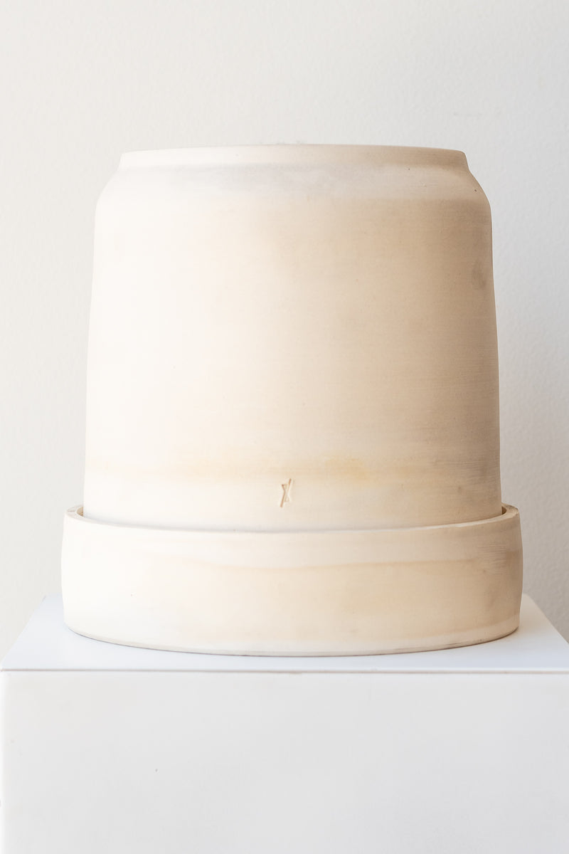 One cream colored stoneware planter sits on a white surface in a white room. The planter is round and tall, with a small logo imprinted in the clay on the bottom. The planter also sits on a round drainage tray. It is photographed straight on.