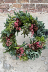 Evergreen single sided wreath with pine cones and pink pepper berries against a grey wall. 