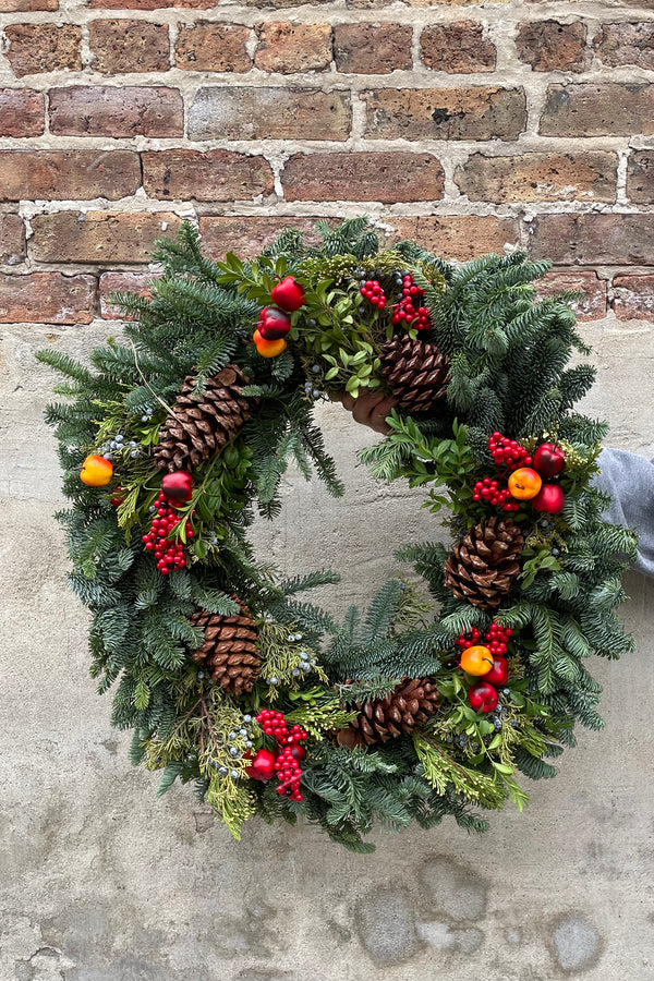 Royal Fruit wreath with pine cones and faux bright fruit  against a concrete wall.