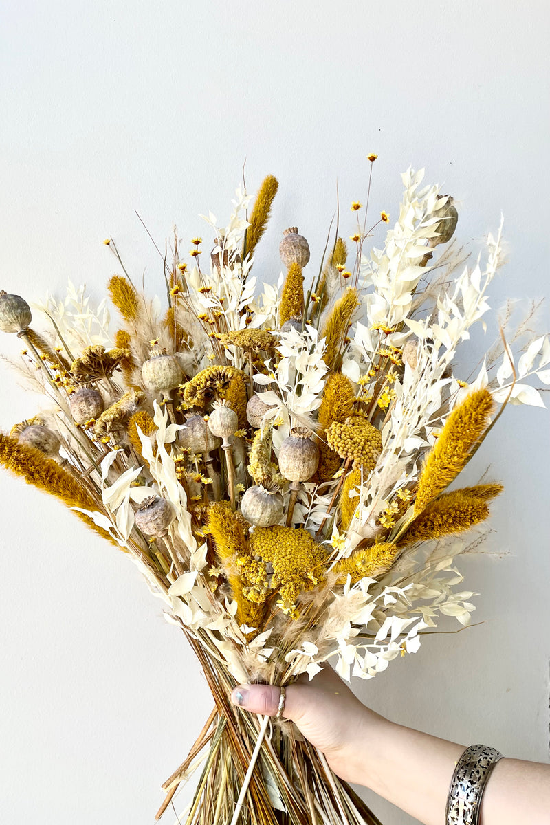 Lushes dried floral arrangement with bright yellows, tans and whites. Hand holding arrangement in front of white wall.